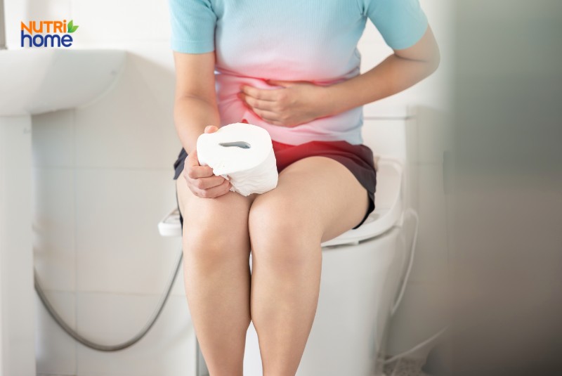Người phụ nữ đau bụng, cầm giấy vệ sinh khi ngồi trên bồn cầu trong nhà vệ sinh