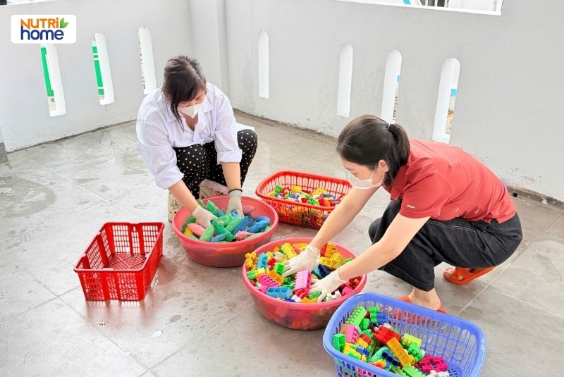 Hai giáo viên ngồi rửa đồ chơi xếp hình trong chậu xà phòng ở sân trường