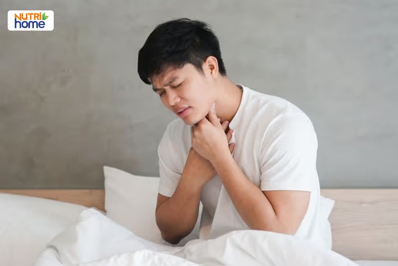 Nam giới ngồi trên giường, đưa tay ôm cổ vì đau rát vùng họng