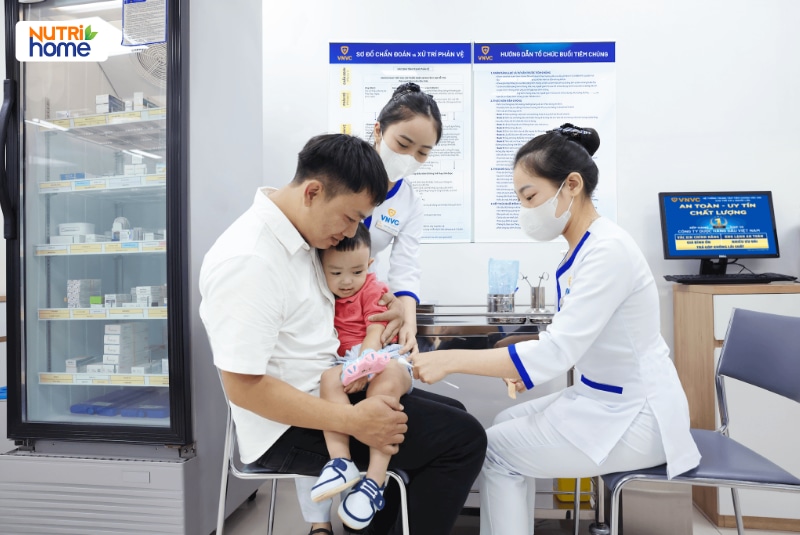 Điều dưỡng VNVC tiêm vắc xin vào đùi bé trai ngồi trong lòng bố
