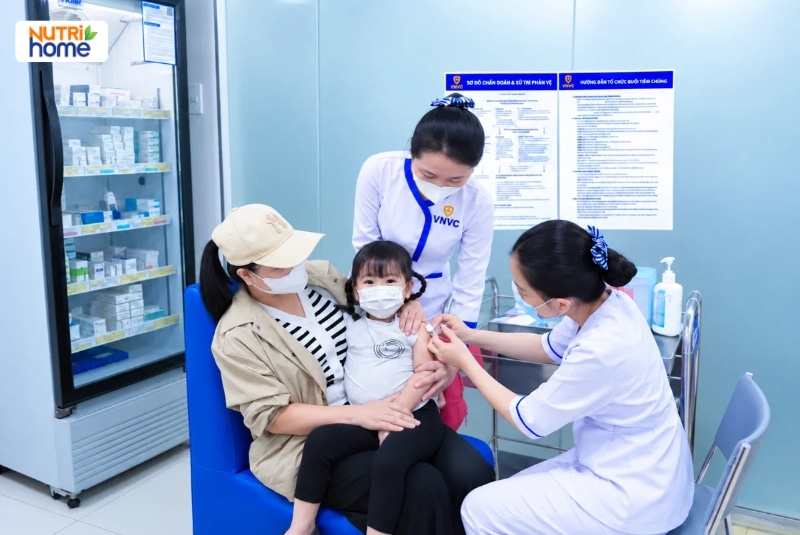 Bé gái ngồi lòng mẹ được điều dưỡng VNVC tiêm vắc xin vào bắp tay.
